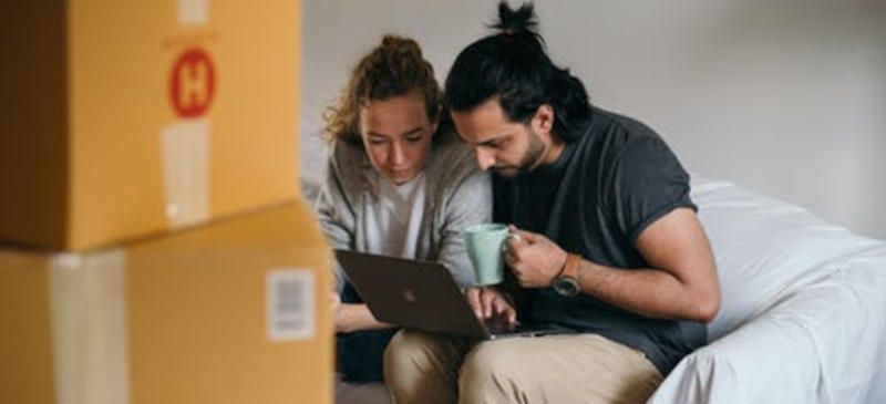 Couple sitting down using laptop together surrounded by moving home boxes